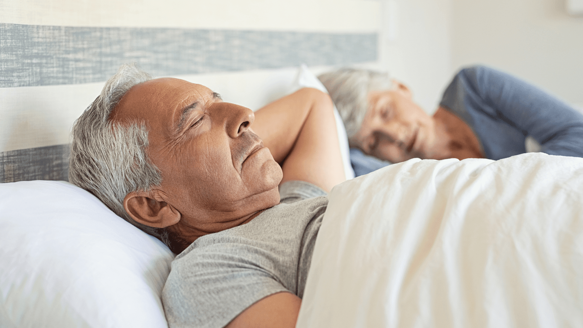 An older couple asleep in bed under a white duvet, the man nearest the camera lying on his back