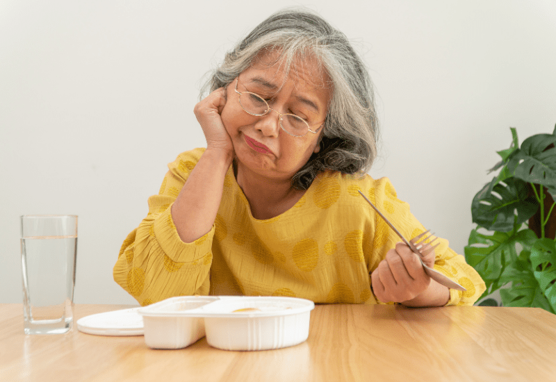 An older woman rests her chin on her hand, fork in the other, looking without appetite at a small dish of food