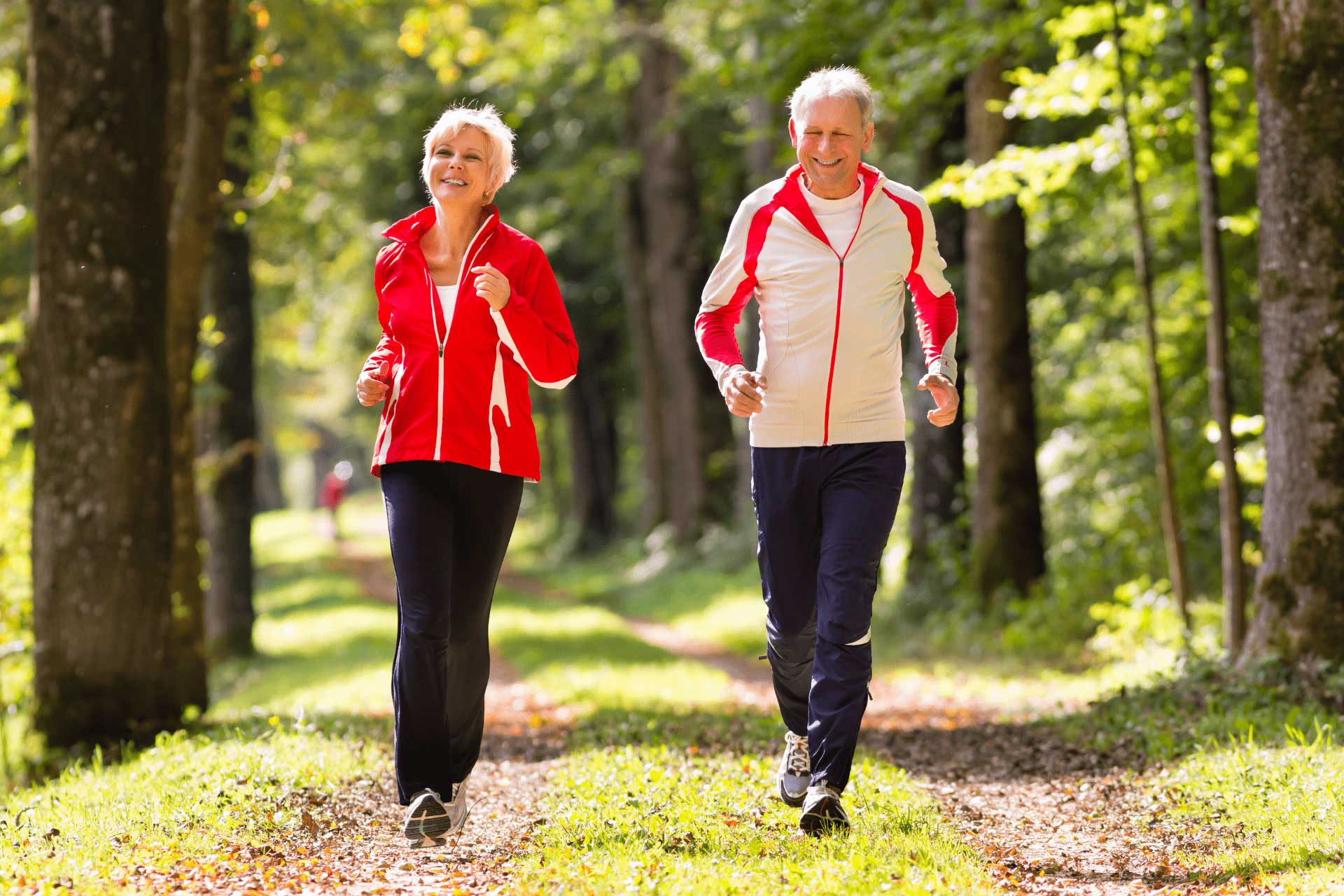 An older couple jogging side by side along a woodland path in matching red and white jackets